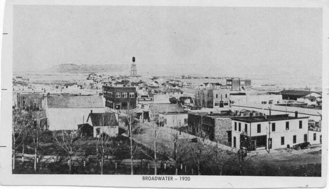 A black-and-white photograph of Broadwater in 1920, showcasing a small town with various buildings, a prominent water tower, and a backdrop of open plains and hills. Leafless trees line the foreground, adding a sense of depth to the scene.
