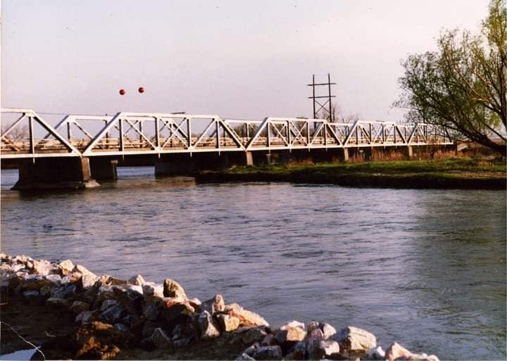 A white metal bridge spans a calm river, with rocks lining the riverbank in the foreground. Two red spheres are suspended above the bridge, and a tall utility pole stands nearby. A tree with sparse foliage is visible to the right, under a clear sky.
