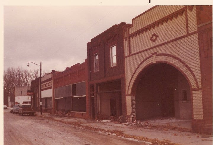 A street scene shows a row of old brick buildings with a large arched entrance and some structural damage. The road is unpaved, and a few parked cars are visible. The sky is overcast, adding to the desolate atmosphere.